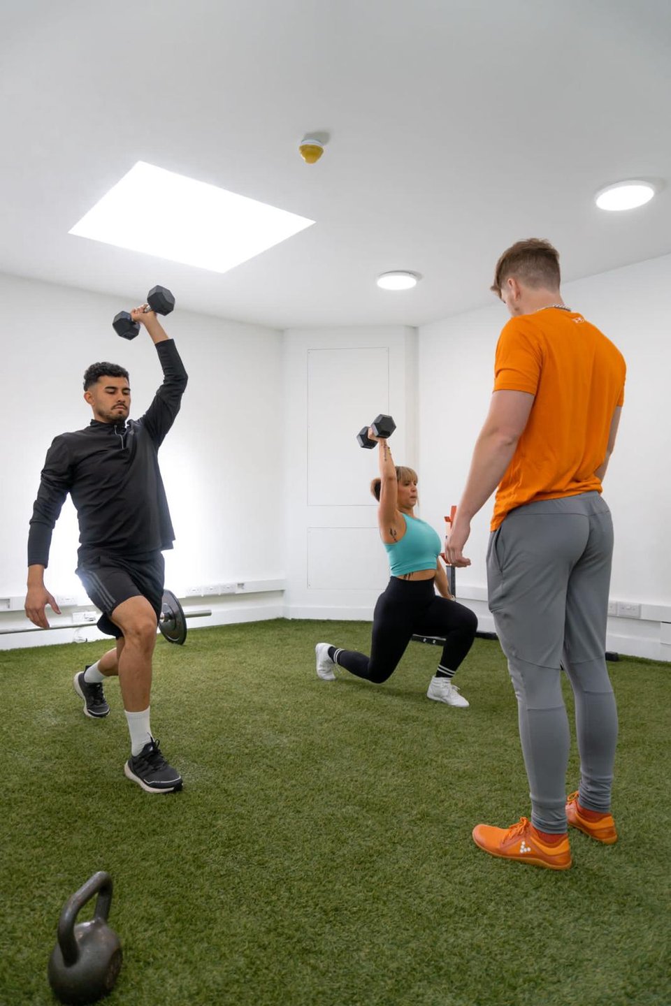Victor coaching two clients through a kneeling overhead dumbbell press on the studio turf