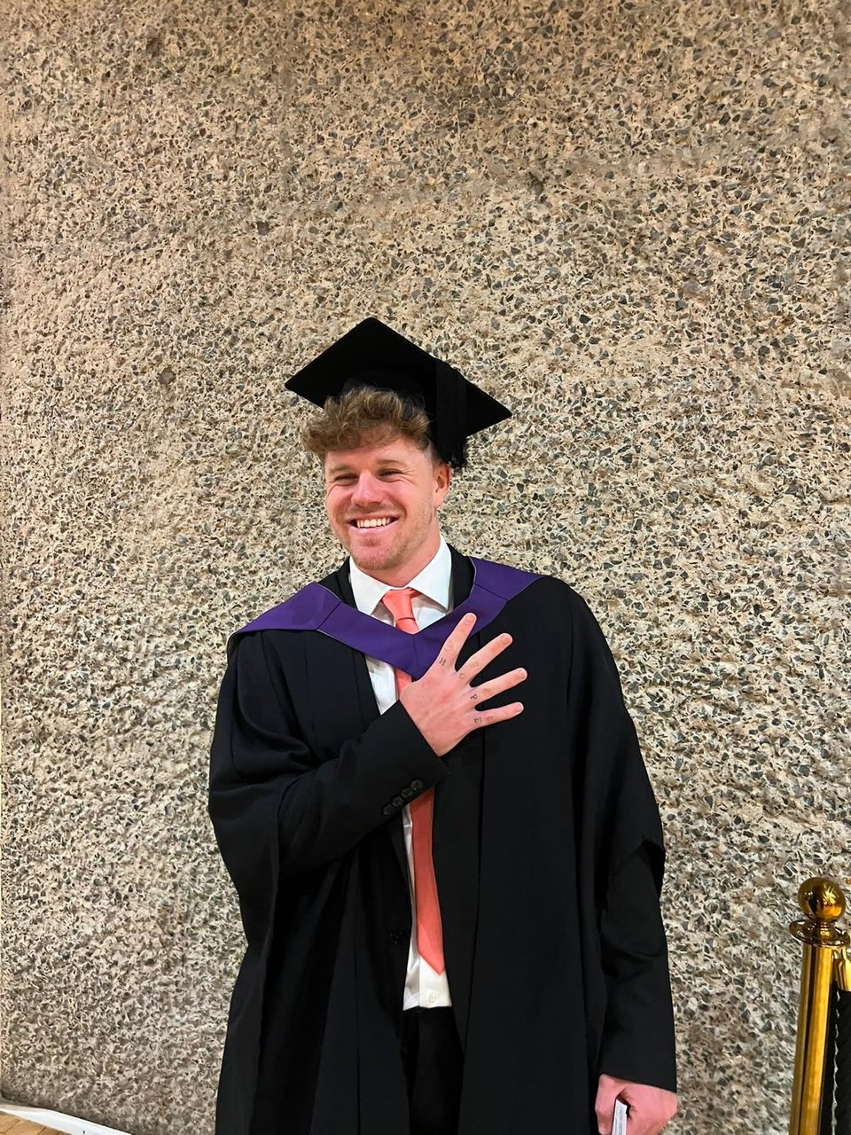 Victor Rambla in his graduation gown, hand on heart, after his First-Class Sports Therapy degree